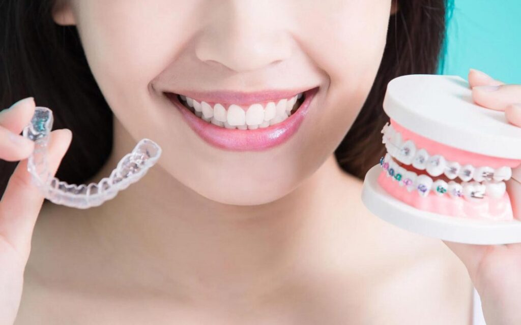 A smiling woman holds a clear Invisalign aligner and ceramic braces, comparing treatment options.